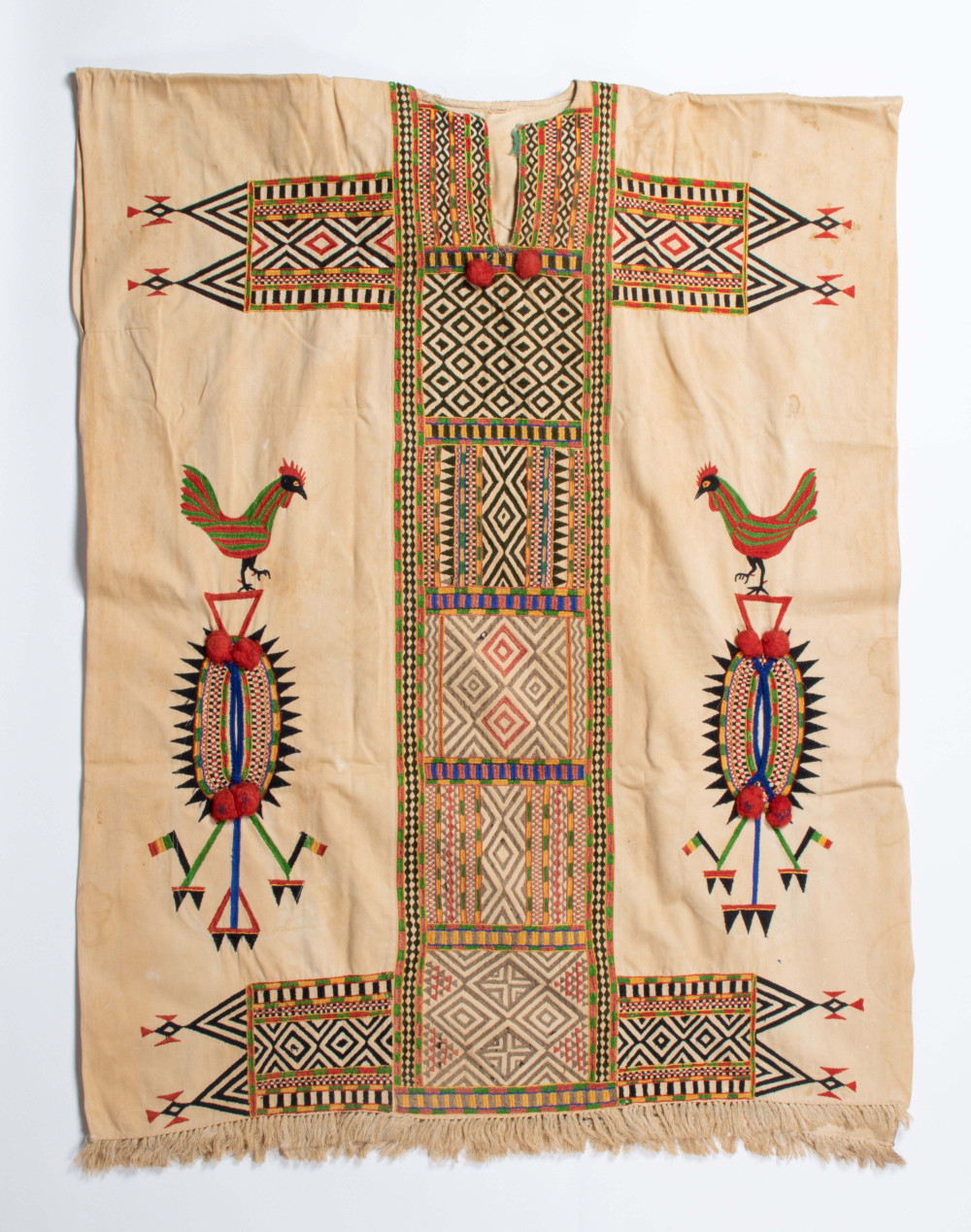 . Beige boubou uit Djennéss (1971-2006), versierd met machinaal borduurwerk, franjes en pompons. De meeste borduursels zijn abstract (strepen, driehoeken en ruiten), met twee rode en groene hanen aan de zijkanten. De zak is aan beide kanten toegankelijk. (7269-3)
