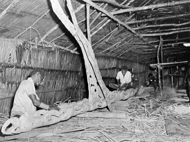 Het vervaardigen van een bisj-paal door houtsnijders, Asmat- regentschap, 1955. Eén steunwortel vormt altijd het uitstekende deel - de tjemen (penis). Fotograaf onbekend. (TM-10010136)